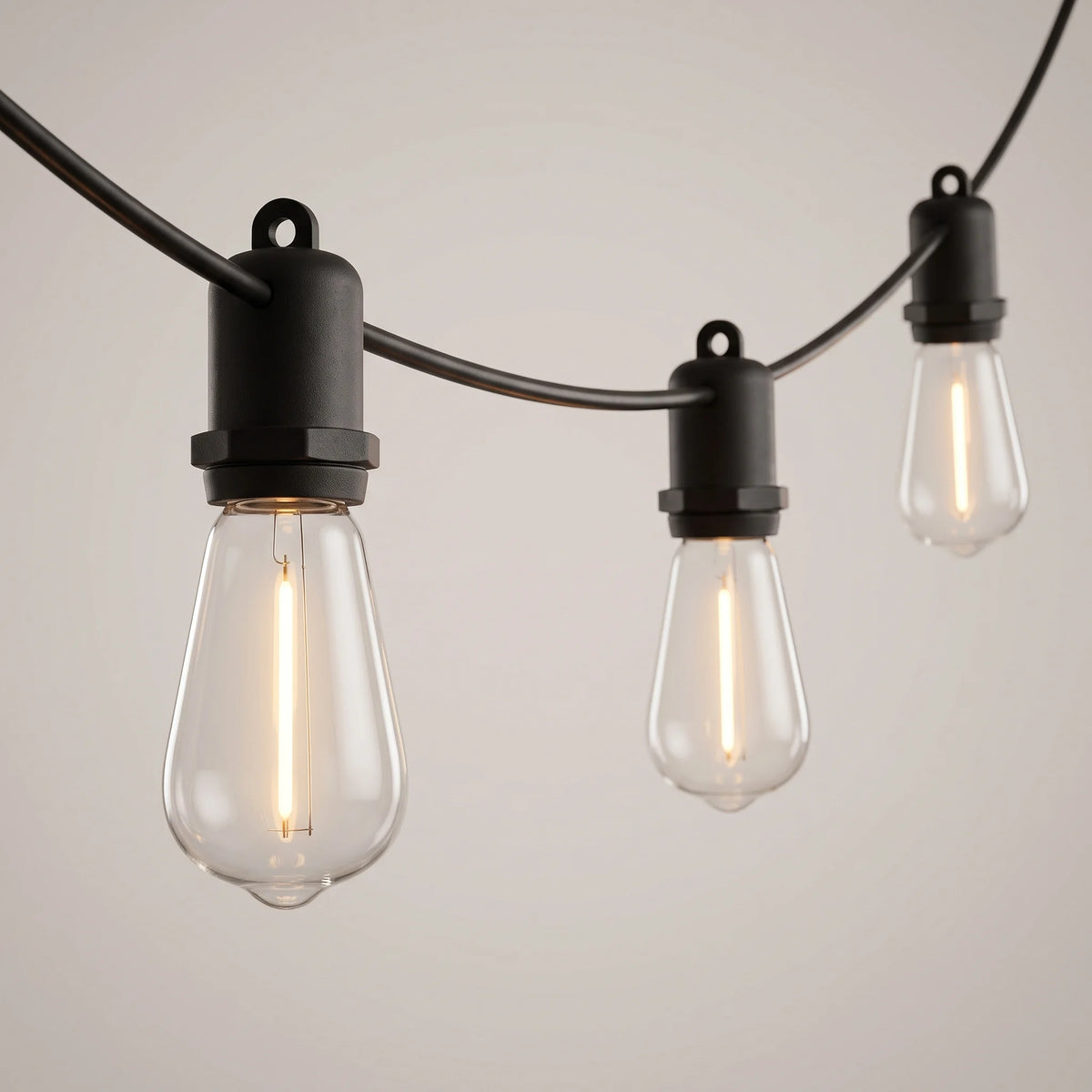 Three Edison bulbs in black weatherproof sockets lit on a black cable against a plain pale background