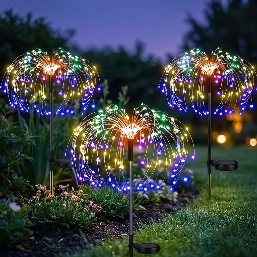 Three solar firework stake lights lit in a garden border at dusk, showing warm white, blue, green and purple LEDs