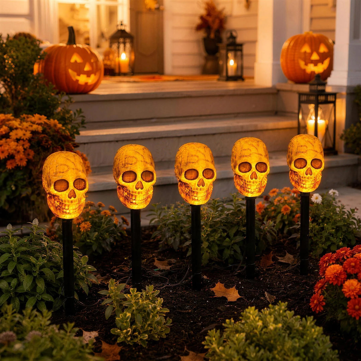 Five lit skull stake lights in a flower border beside porch steps at dusk, with jack-o-lanterns behind