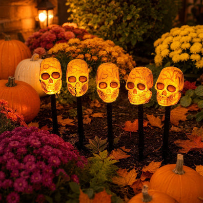 Close view of five lit skull stakes among autumn flowers and fallen leaves, glowing warm orange
