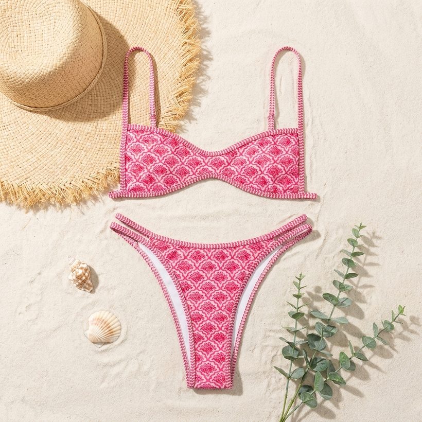 Pink scalloped-print bikini top and bottoms laid flat on sand with a straw hat and shells
