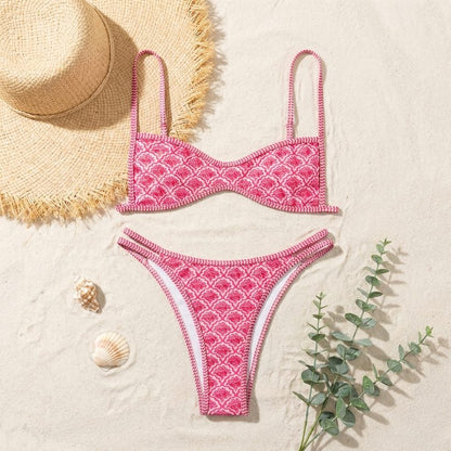 Pink scalloped-print bikini top and bottoms laid flat on sand with a straw hat and shells