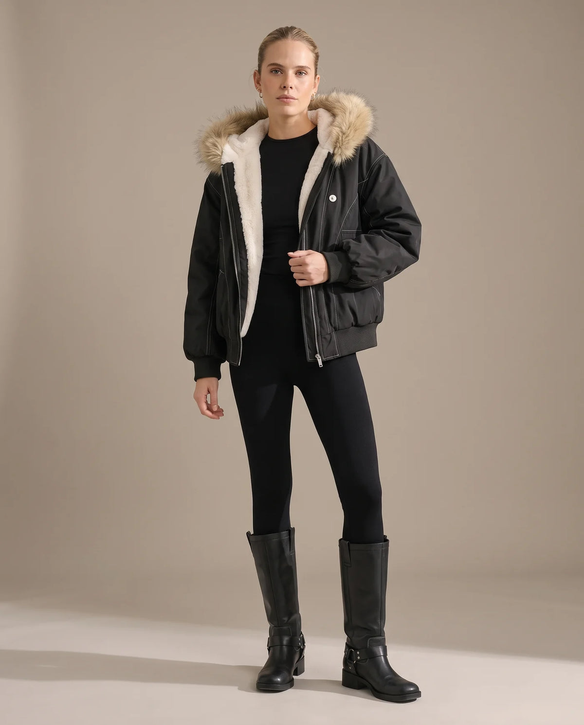 A woman wearing the black bomber jacket open over black leggings and knee-high boots