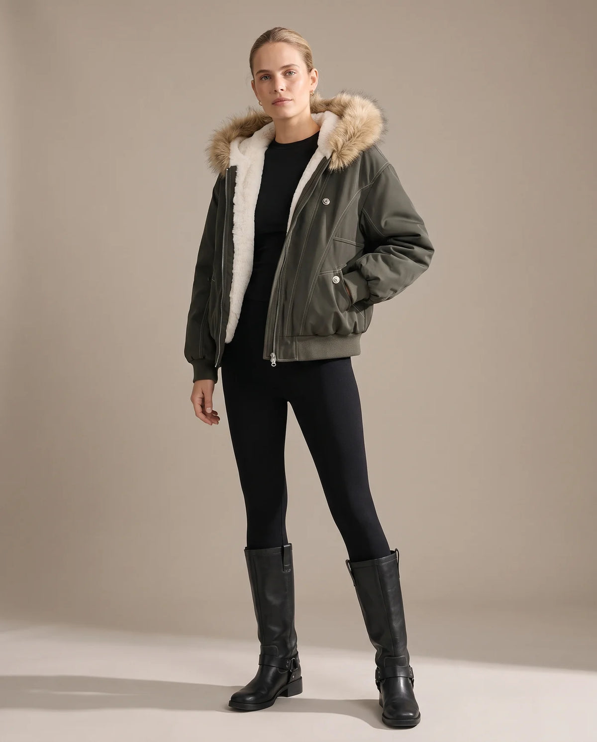 A woman wearing the army green bomber jacket open over black leggings and knee-high boots